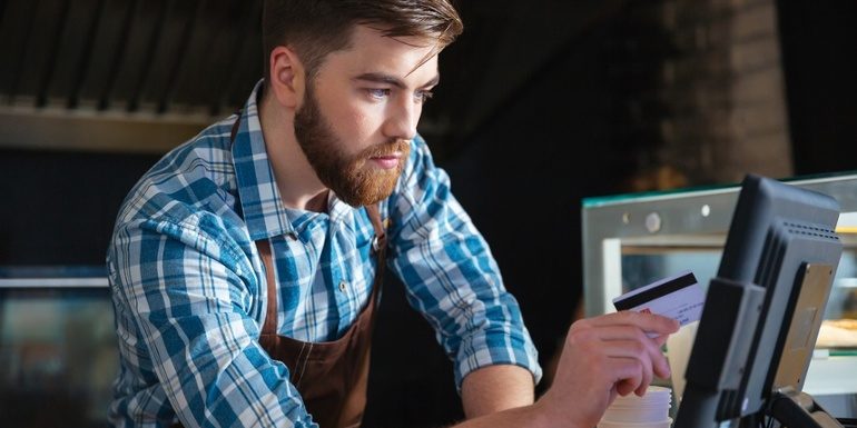 Handsome concentrated bearded waiter swiping credit card through the computer terminal in cafe